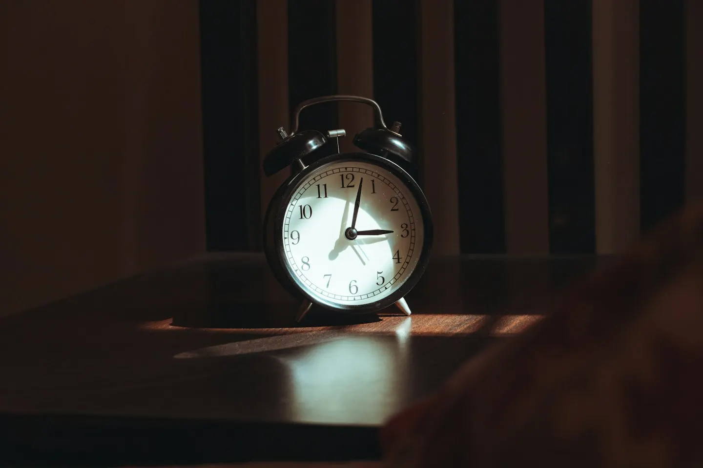 A black twin-bell alarm clock on a wooden bedside table, lit by a thin strip of light in a dark room.