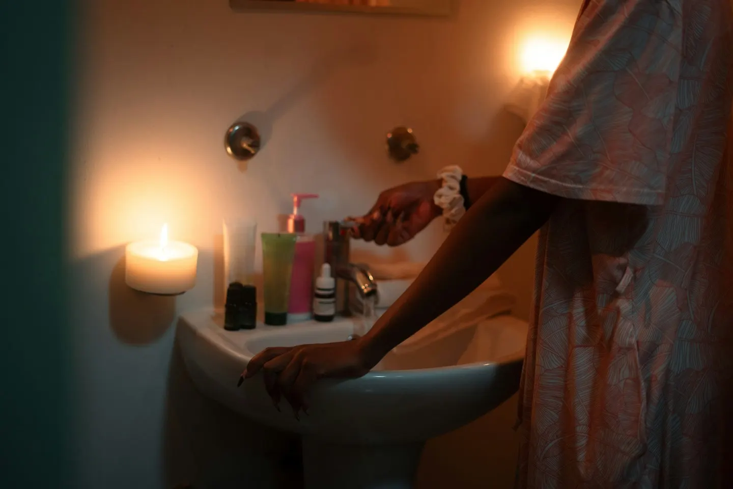 A person in a patterned nightdress standing at a bathroom sink at night, lit by a candle and a small wall lamp, one hand on the basin and the other on the tap.