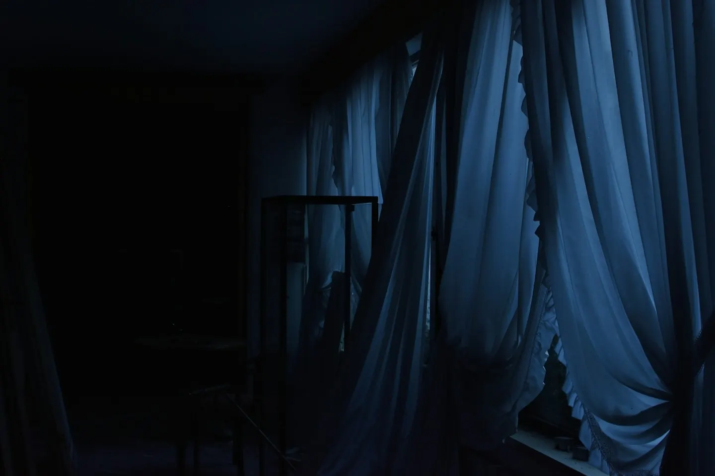 A dark bedroom at night with pale sheer curtains lit faintly blue from the window and the outline of a chair in the shadows.