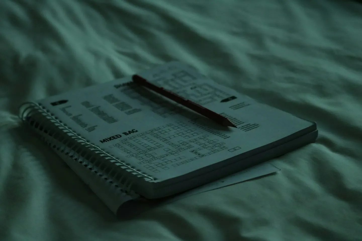A spiral-bound puzzle book with a red pencil lying on rumpled bed sheets, in dim green light.