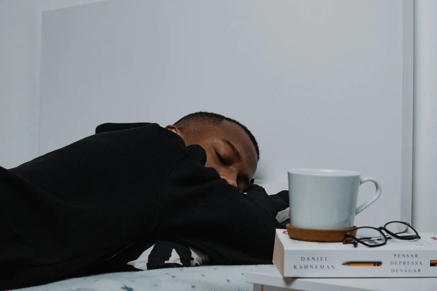 A young man in a black sweatshirt asleep face down on a bed, next to a bedside table with a white mug and glasses resting on a book.