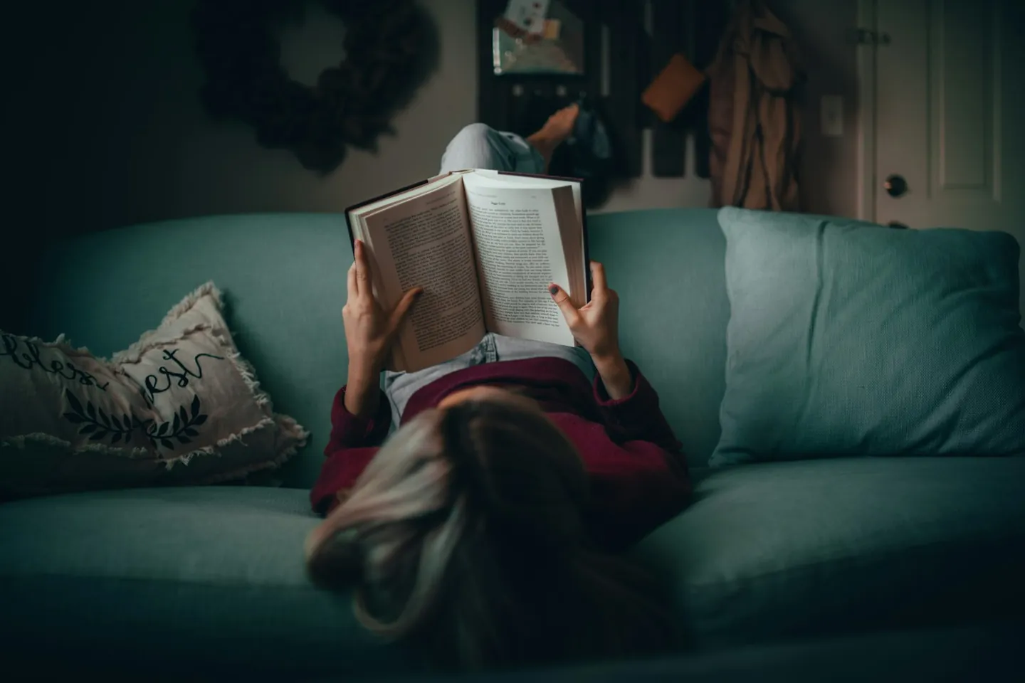 A woman in a red shirt lying on a sofa in a dim living room, holding an open book above her face.