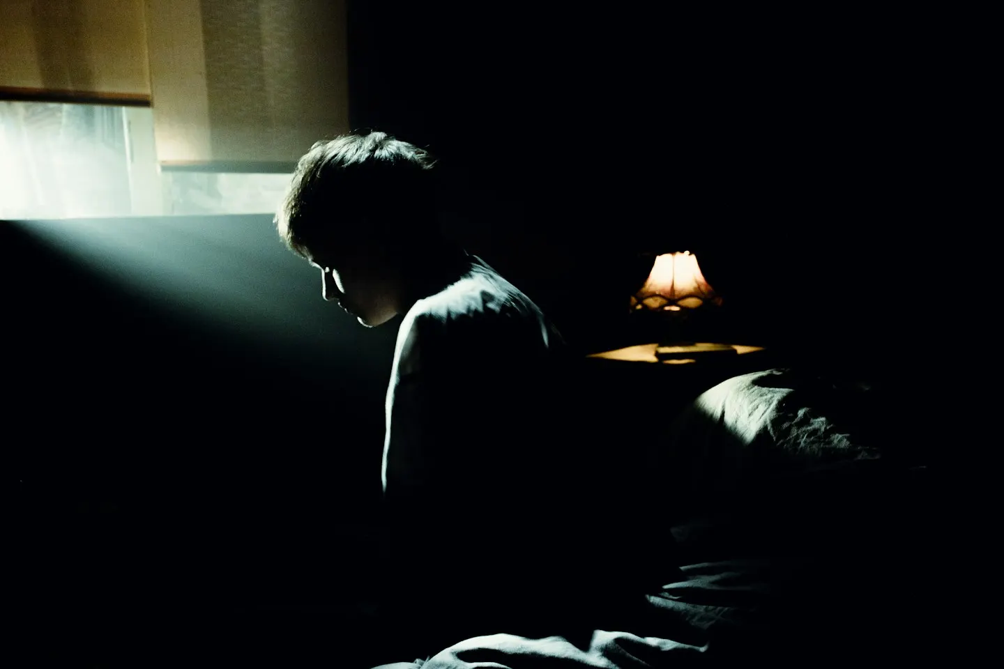 A person sitting hunched on the edge of a bed in a dark room, lit by a thin beam of light from a window, with a small lamp glowing on the bedside table behind.