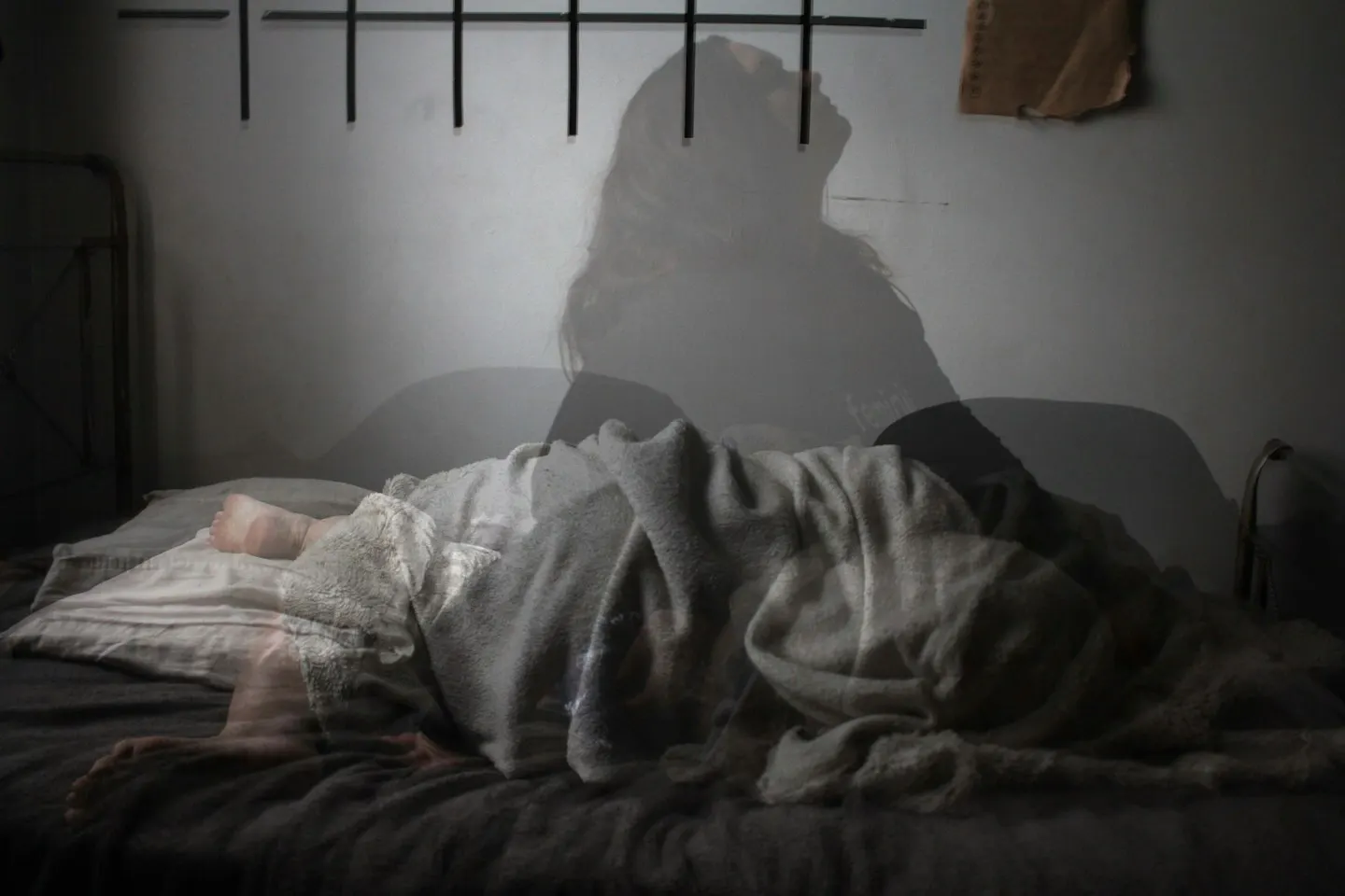A double-exposure photo of a woman lying curled up under grey blankets in a dim bedroom, with a faint shadow of a person sitting up against the wall above her.