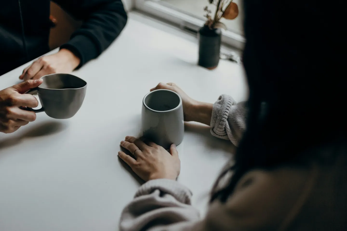 Two people sitting across a white table, each holding a grey mug, seen from above with their faces out of frame.