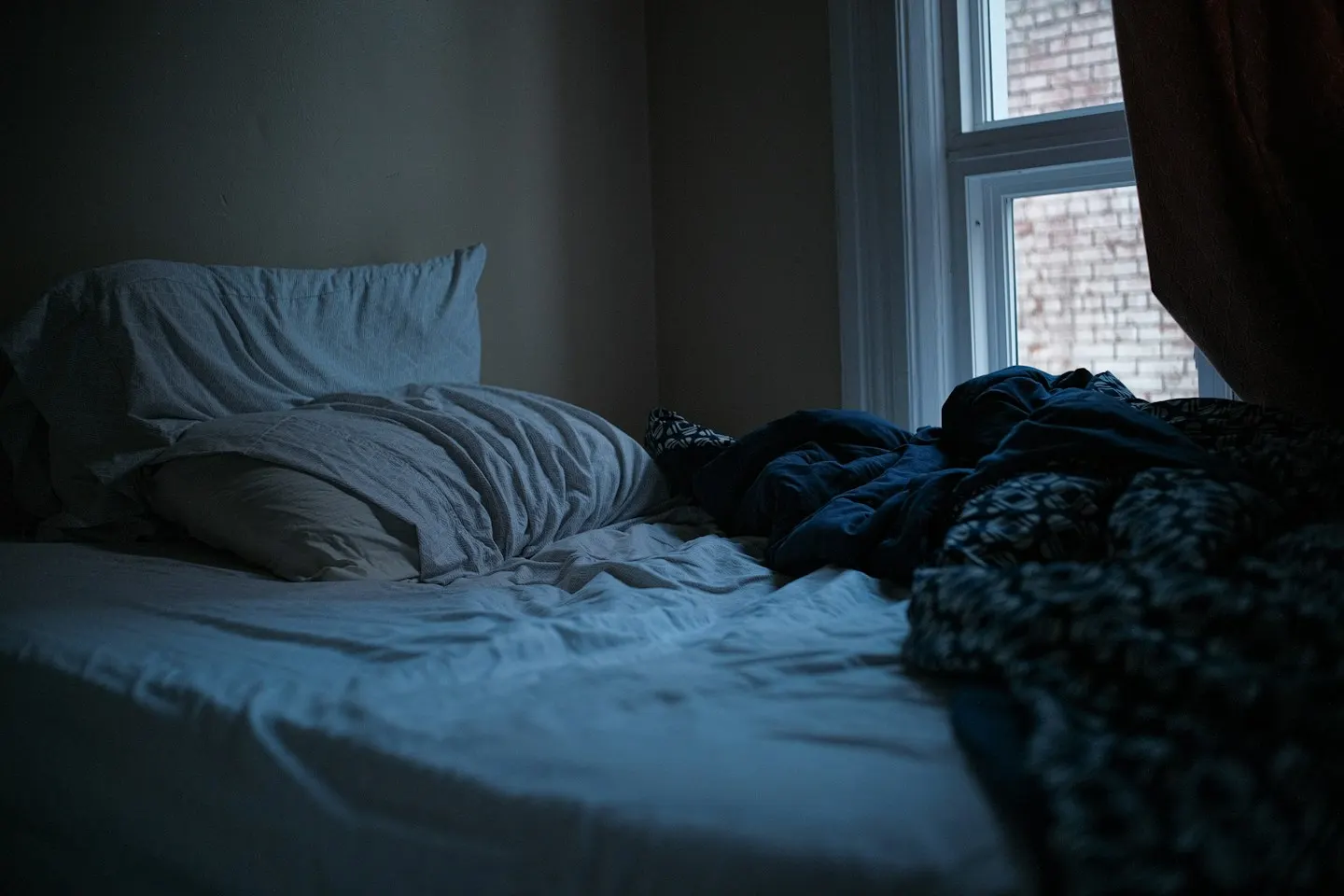 An unmade bed with rumpled white sheets and a dark blue duvet in a dim bedroom, next to a window that looks out onto a brick wall.