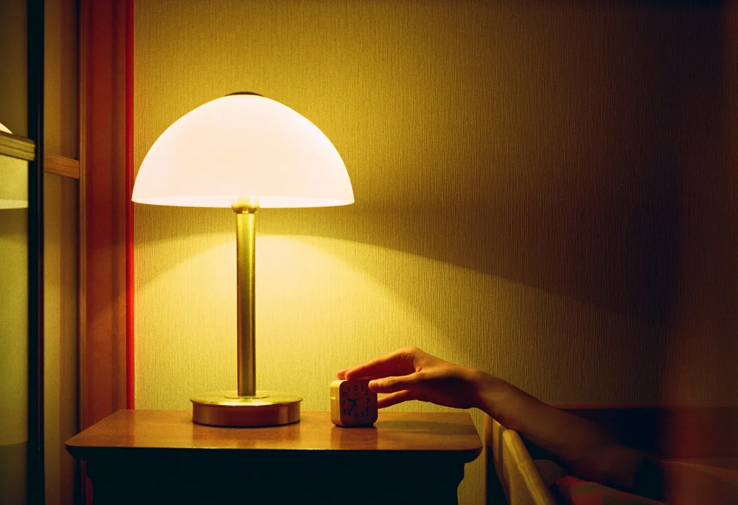A hand reaching from a bed to a small alarm clock on a wooden nightstand, next to a lit bedside lamp in a dim room.