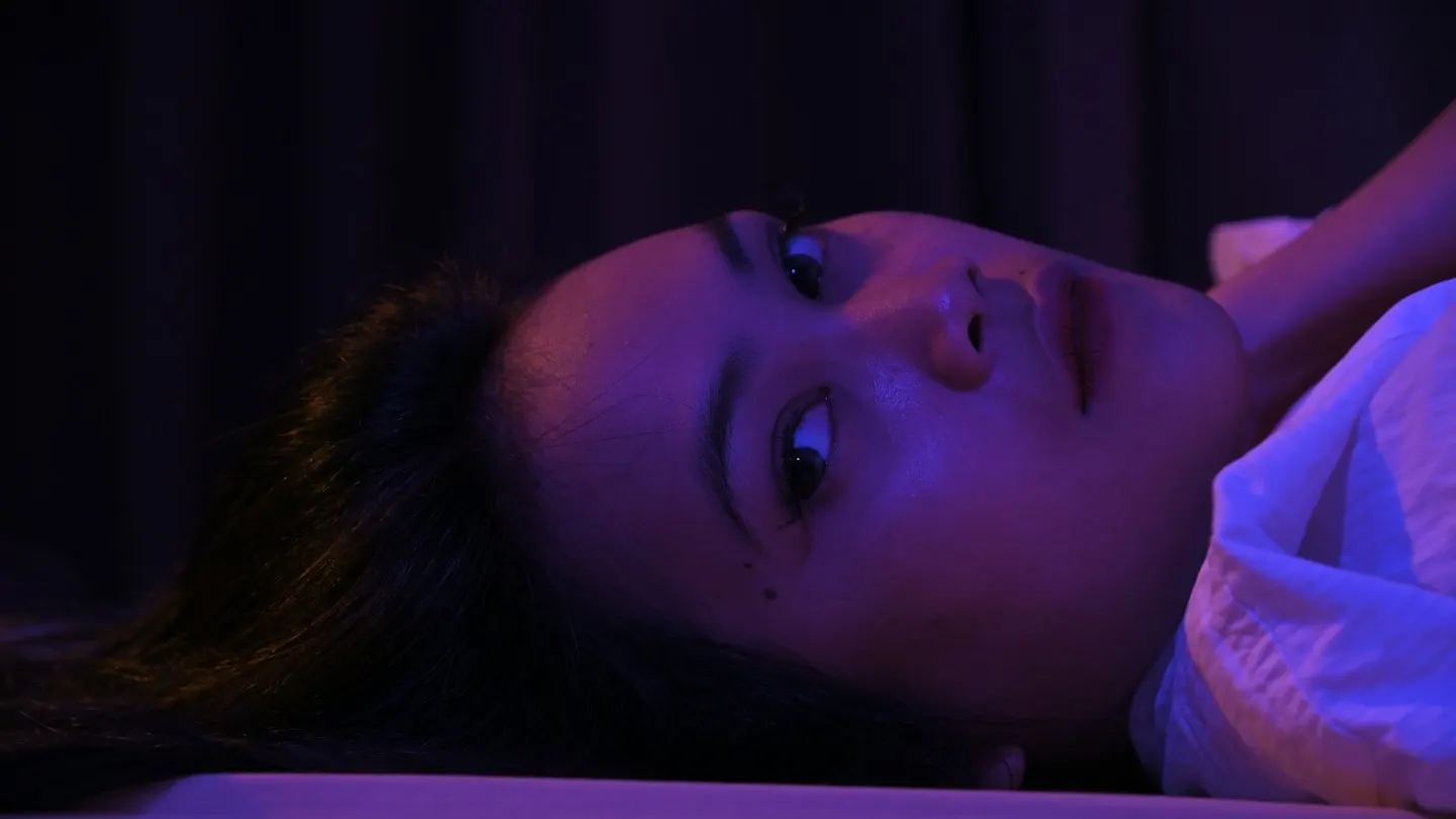 A woman lying on her back in a dark room lit in purple and blue, her eyes open and looking to the side.