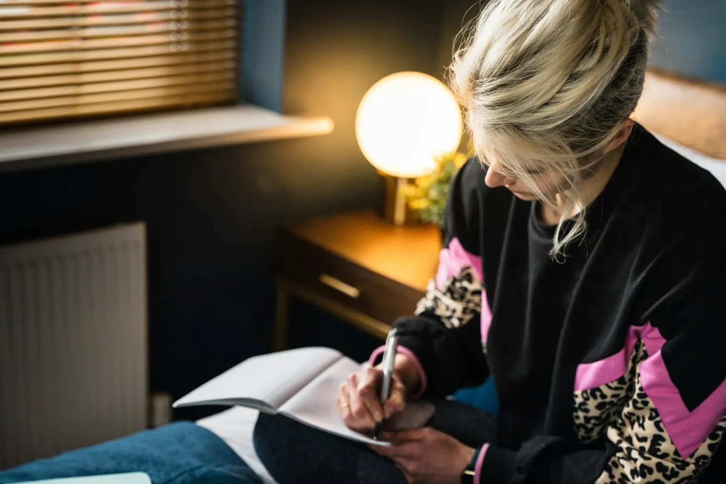 A woman with blond hair sitting on a bed, writing in an open notebook next to a lit bedside lamp.